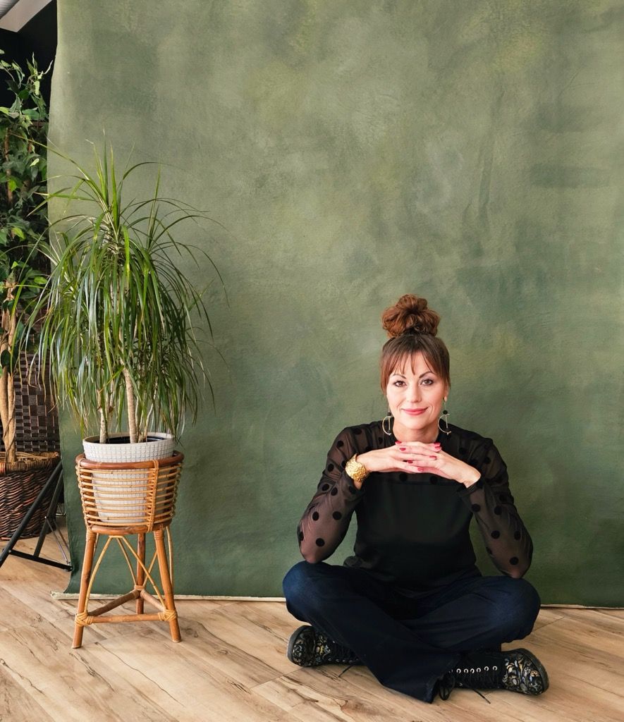A person in a black outfit sits cross-legged on a wooden floor next to a potted plant against a sage green wall.