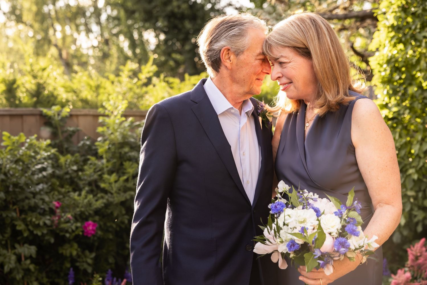 A couple poses together in a garden setting with a bouquet of purple and blue flowers.