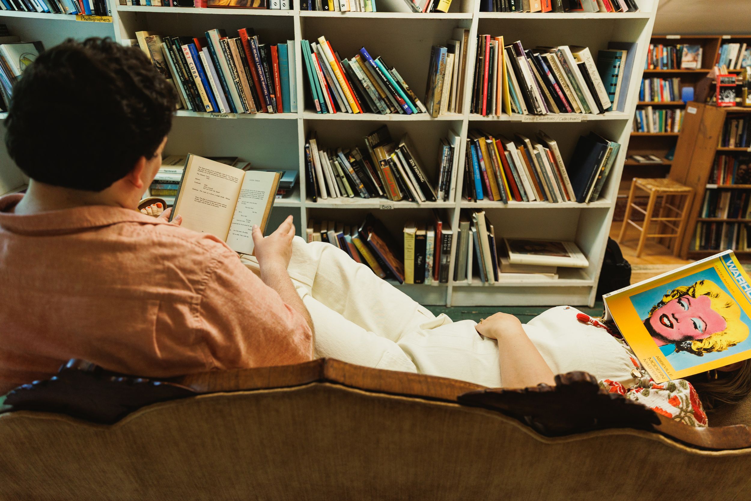 Person relaxing on couch in library reading books with colorful children's book visible.