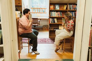 Two individuals sit and chat in a book-filled study with wooden chairs and bookshelves.