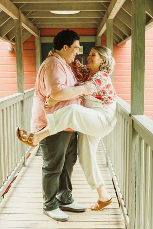 Couple sharing playful moment on wooden porch with coral colored building in background.