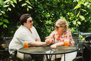Two people engage in conversation while seated at a black metal patio table with orange beverages.