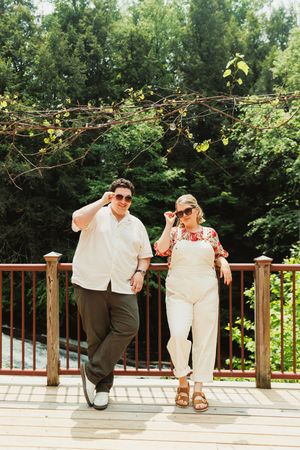 Two people relax and pose playfully on a wooden deck railing surrounded by lush green foliage on a sunny day.