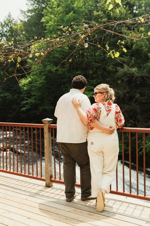 A couple standing together on a wooden deck overlooking nature during a sunny day.