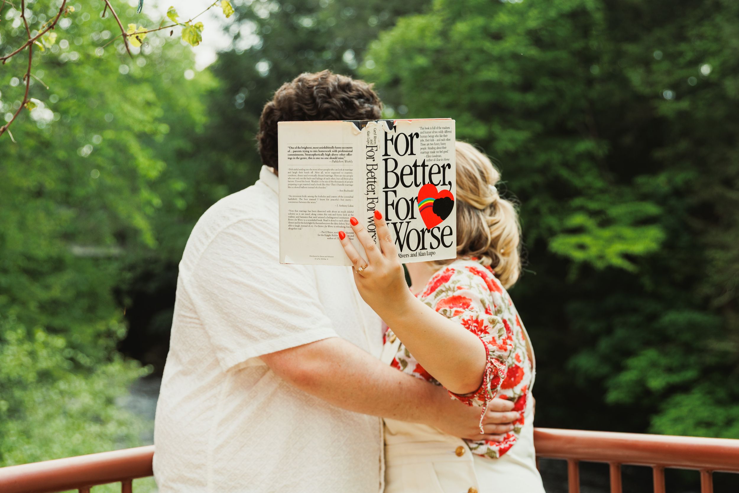 Two people embracing while holding a book on an outdoor wooden deck surrounded by greenery.