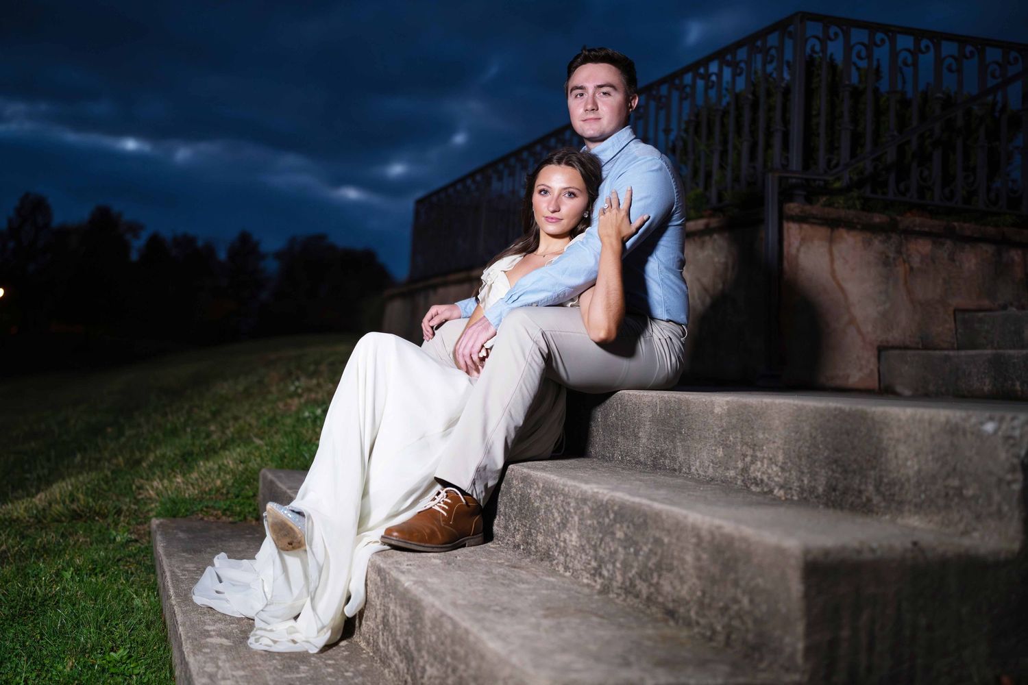 A couple in formal wedding attire poses on concrete steps at dusk with dramatic stormy skies in the background.