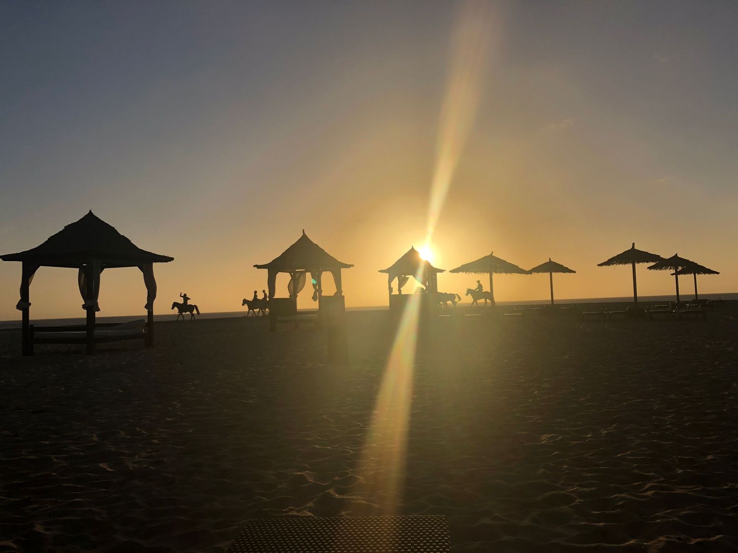 Silhouetted beach cabanas and umbrellas against a bright sunburst at sunset on a sandy beach.