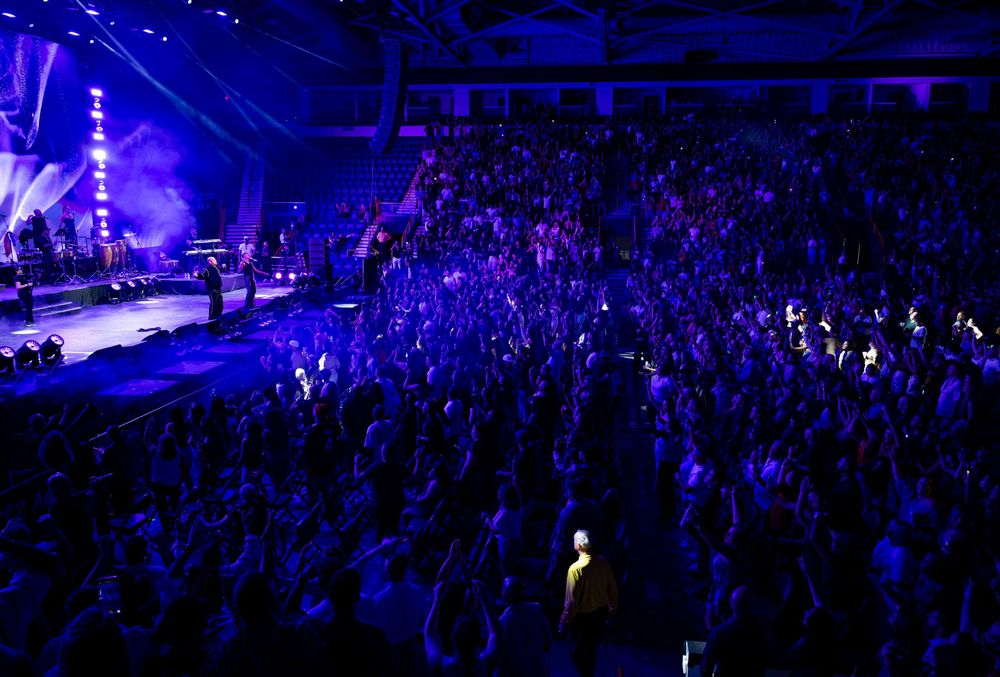 Massive crowd of fans with raised hands fills an arena during a live concert with blue stage lighting.