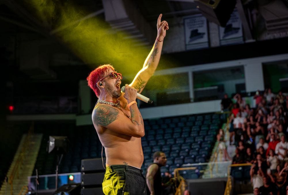 Shirtless tattooed male performer with red hair singing on stage under yellow spotlights at a live concert.
