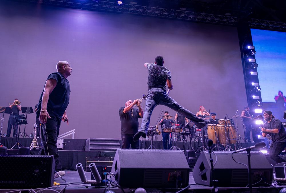 Male performer leaps off stage monitor while another artist watches during an energetic live concert.