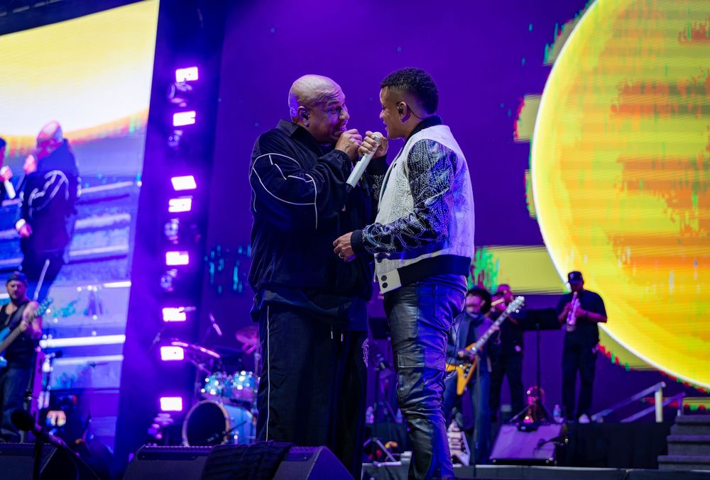 Two male artists sharing a microphone on a colorful concert stage with a large yellow sphere backdrop.
