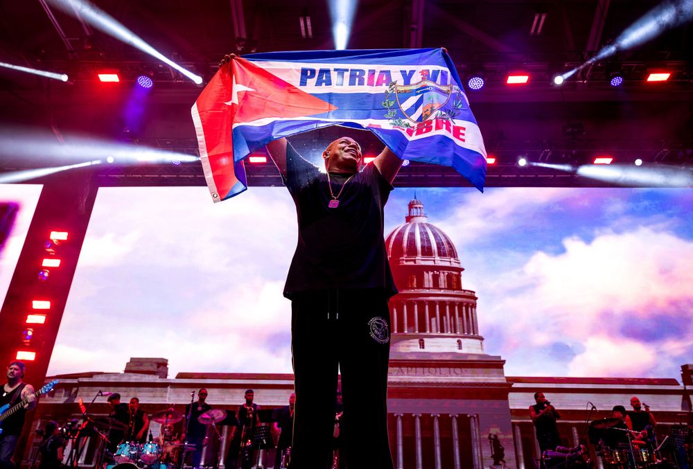 Performer holds a Cuban Patria y Vida flag on a dramatic stage with capitol building backdrop.