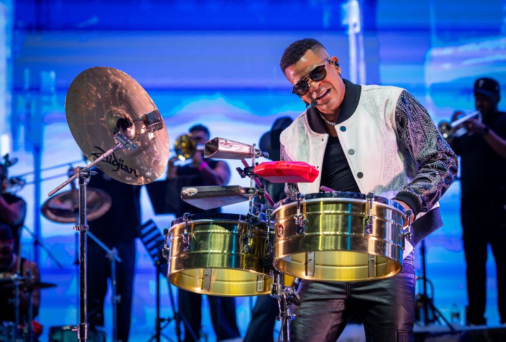 Energetic percussionist playing timbales on a bright concert stage with a full band in the background.