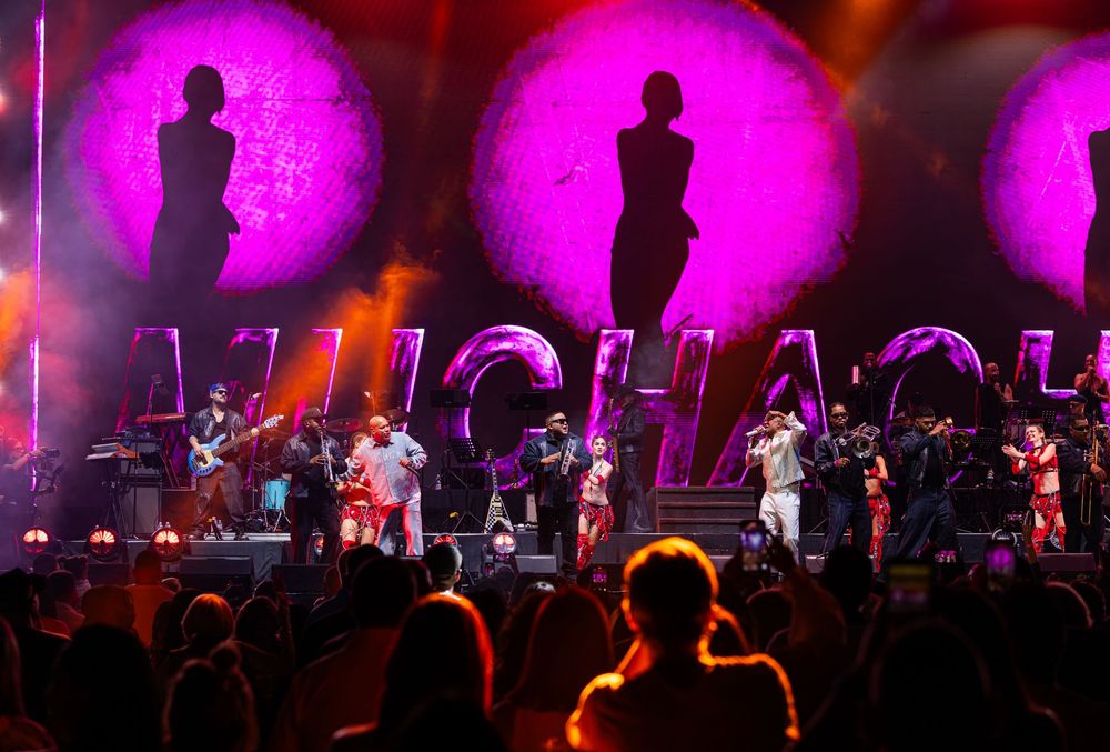 Large band performs on stage with purple lit backdrops displaying silhouettes and the word Muchachos.
