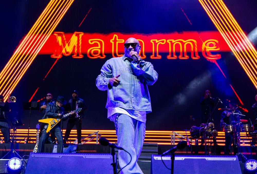 Male rapper performs on stage in front of yellow neon sign reading Matarme with band in background.