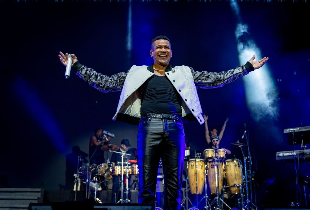 Male performer in white jacket spreads arms wide on a concert stage with percussion instruments behind him.