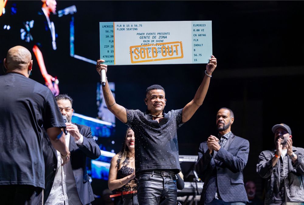 Young male performer holds up large sold-out certificate on stage surrounded by applauding guests.