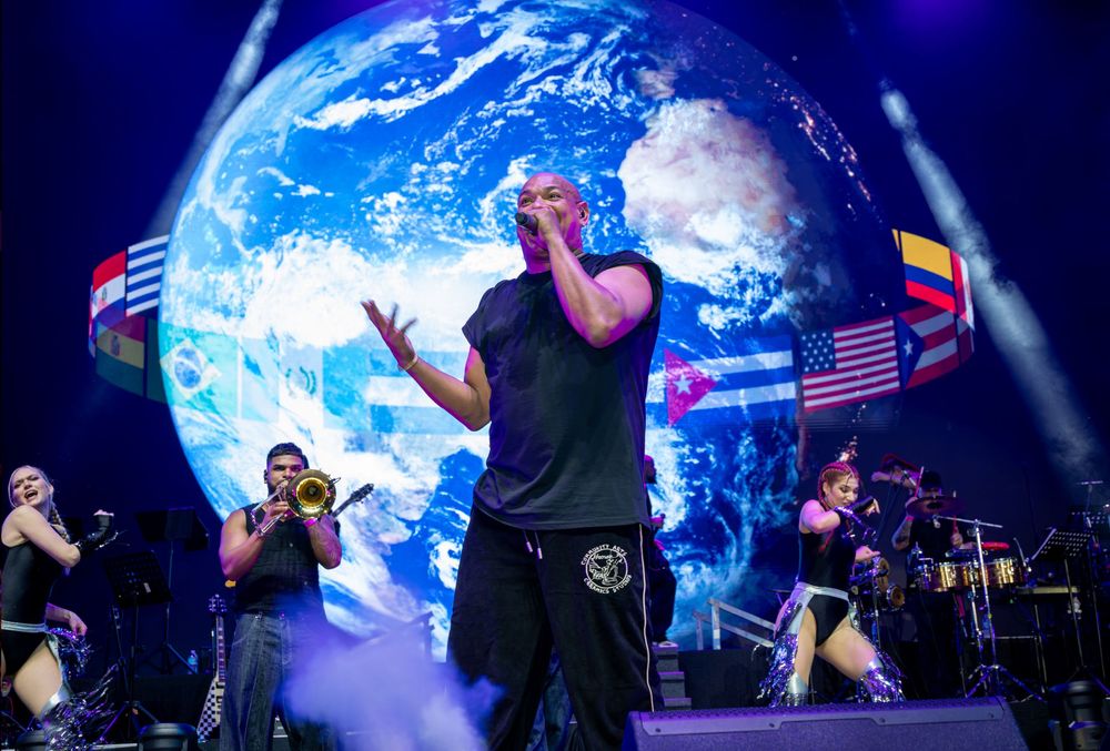 Male singer performs on stage in front of giant globe backdrop with international flags displayed.
