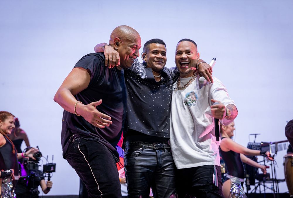 Three male performers share a warm group hug on stage at the end of a live concert.