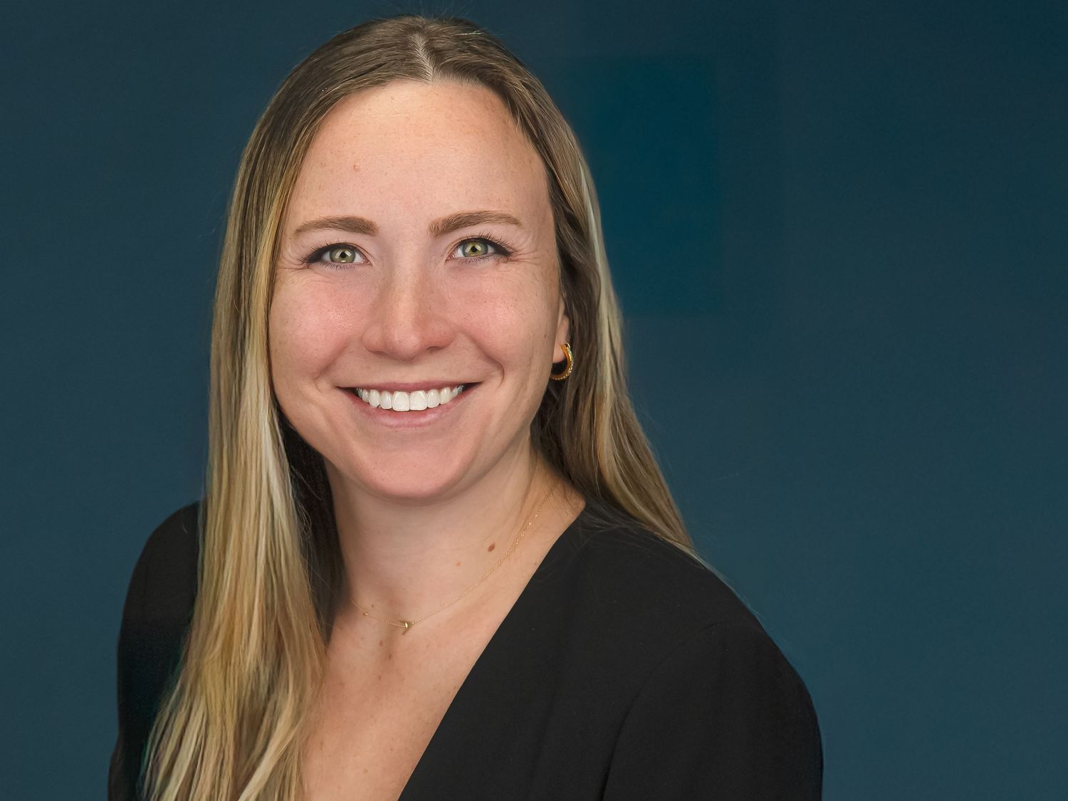 Professional headshot I Greenwich ct of a person with long blonde hair wearing a black top against a dark blue background, smiling warmly.