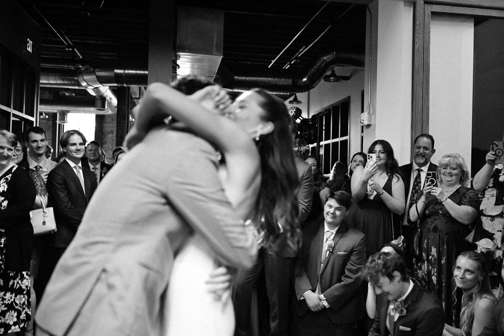 A black and white photograph captures a romantic dip kiss at a wedding celebration as guests look on and smile.