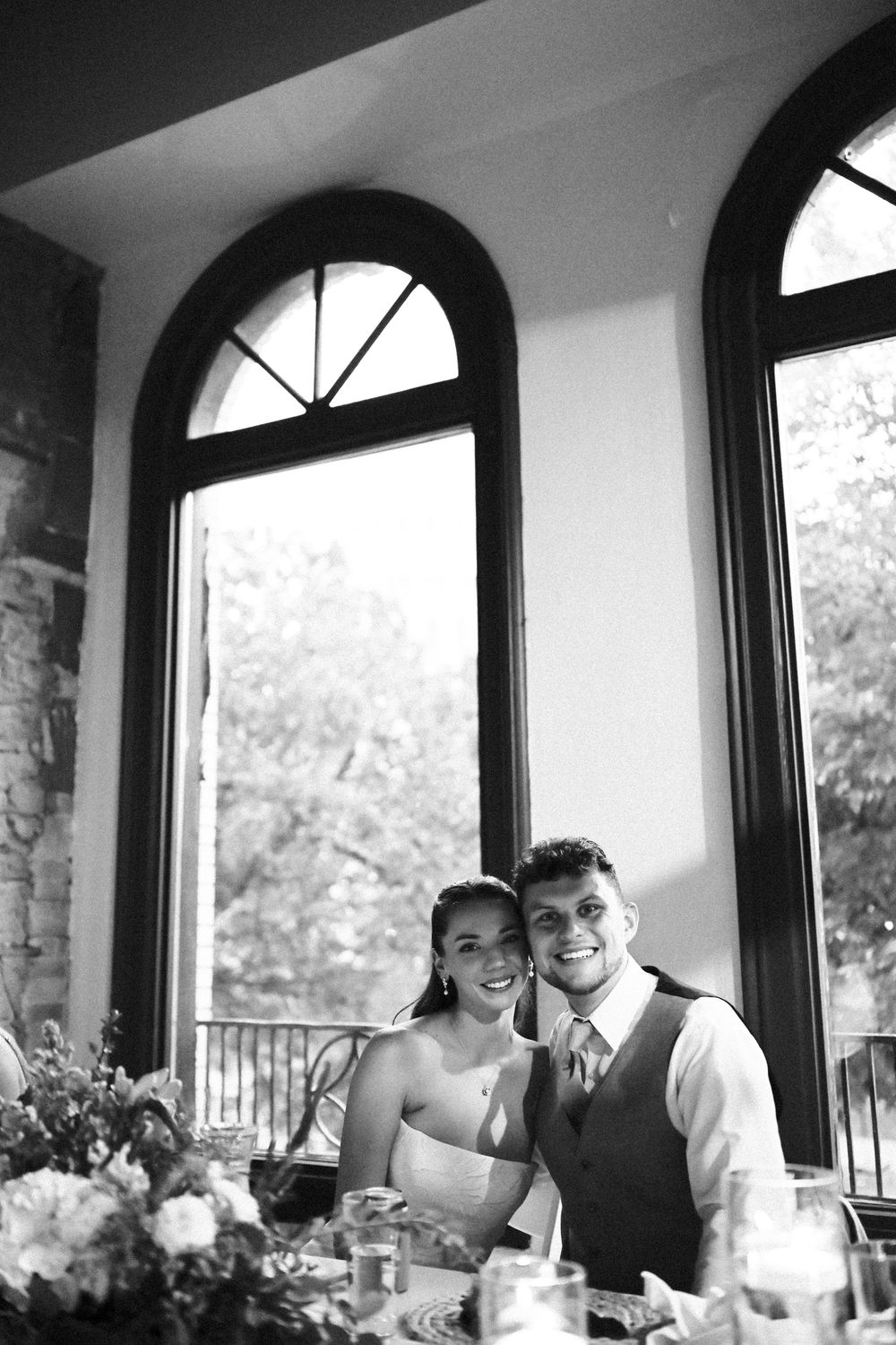 Black and white wedding reception photos show happy couple laughing and embracing at a venue with arched windows.
