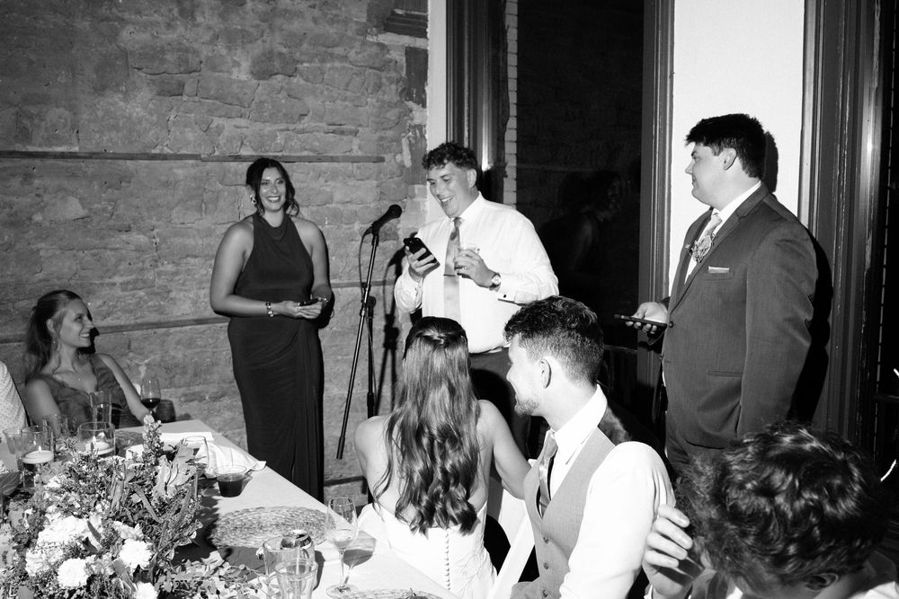 Black and white candid photo of people gathered around a table with floral arrangements at a social event.