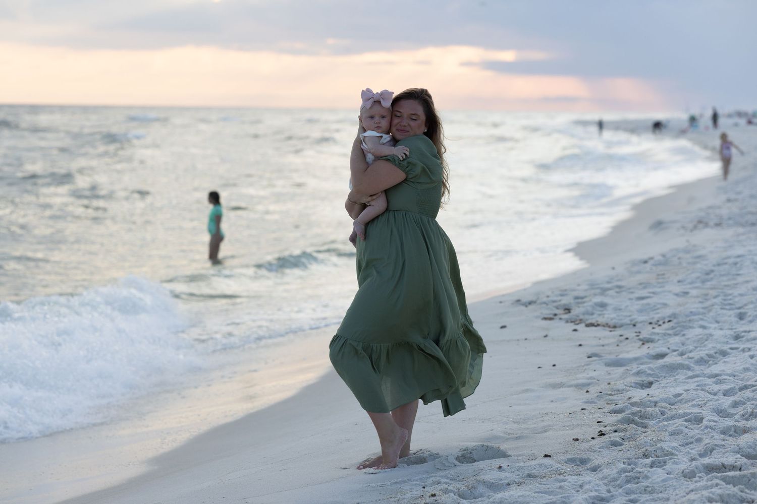 Woman in green dress holds baby on beach at sunset with waves and people in background.