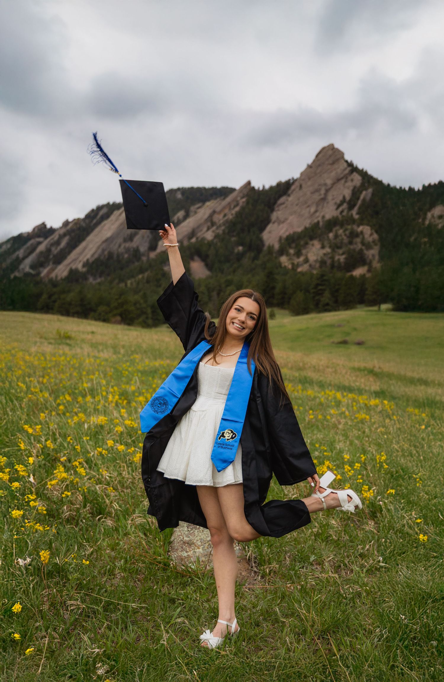 A graduate in cap and gown poses joyfully in a field of yellow wildflowers with the Flatirons rock formation in Boulder behind.