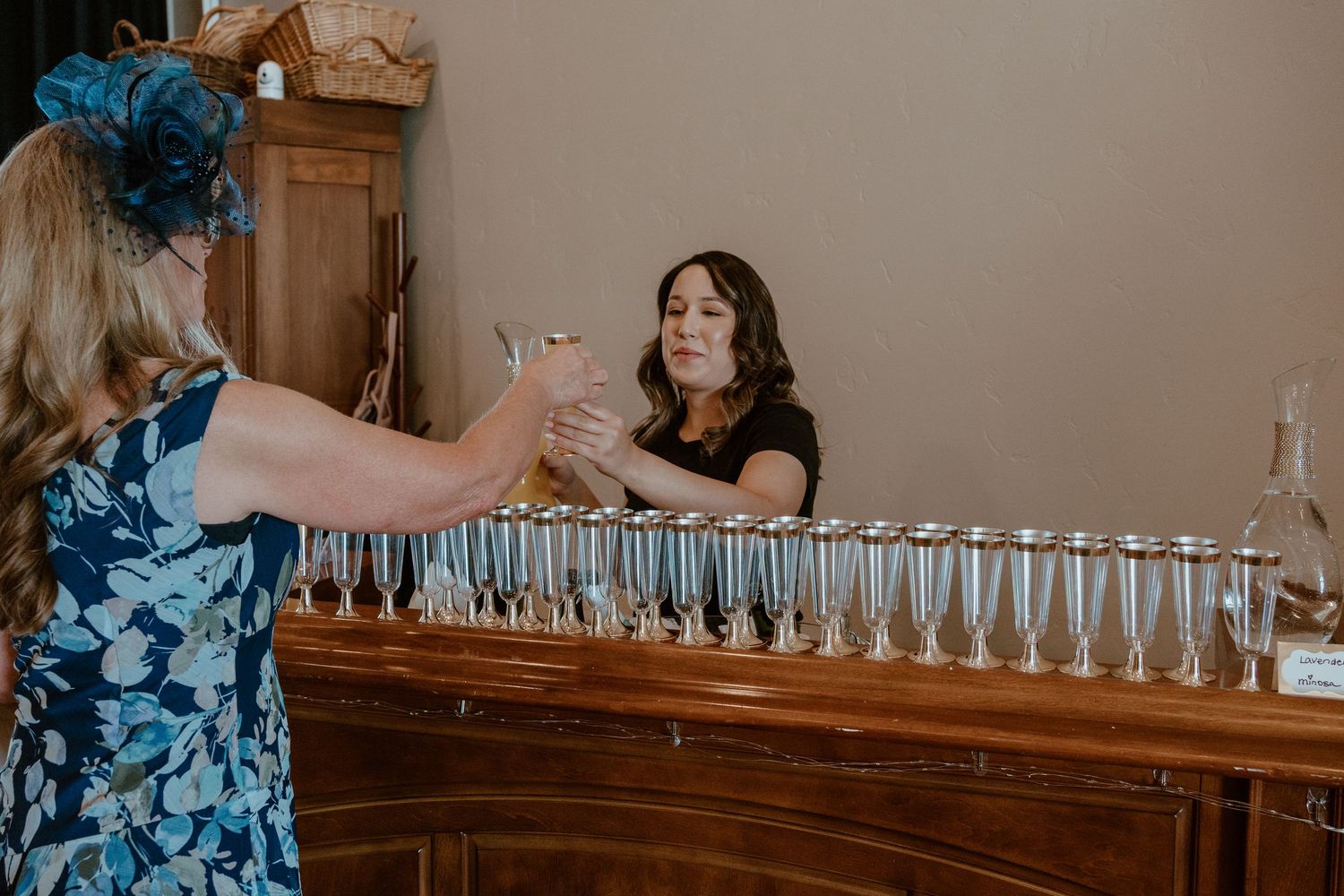 Two women at a bar lined with silver champagne flutes, one handing a glass at an elegant indoor event.