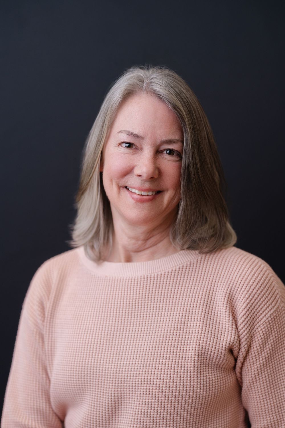 Professional portrait of a woman with silver hair wearing a pink sweater against a dark background.