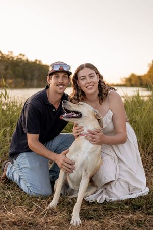 A smiling couple crouches in tall grass at sunset, petting their happy yellow Labrador dog near a river.