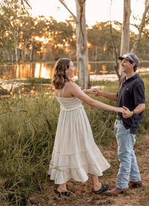 A couple dances and holds hands in a golden sunset forest clearing near a reflective lake surrounded by eucalyptus trees.