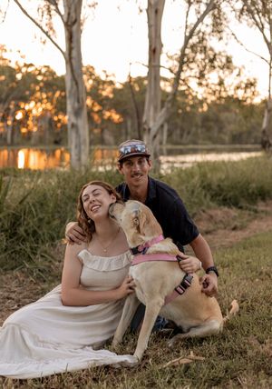 Happy couple sitting on grass with yellow Labrador dog at sunset near a lake.