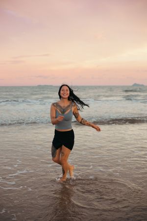A tattooed woman laughs joyfully while running through shallow ocean waves at a pink and purple sunset beach.