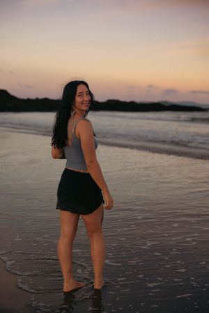 A smiling tattooed woman in a grey crop top and black shorts stands on a beach at dusk looking over her shoulder.