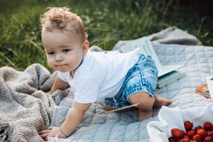 Cute curly-haired baby in white shirt crawling on a blanket outdoors near fresh strawberries.