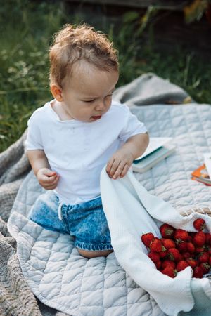 Adorable baby in white shirt and jeans sitting on blanket with bowl of strawberries.