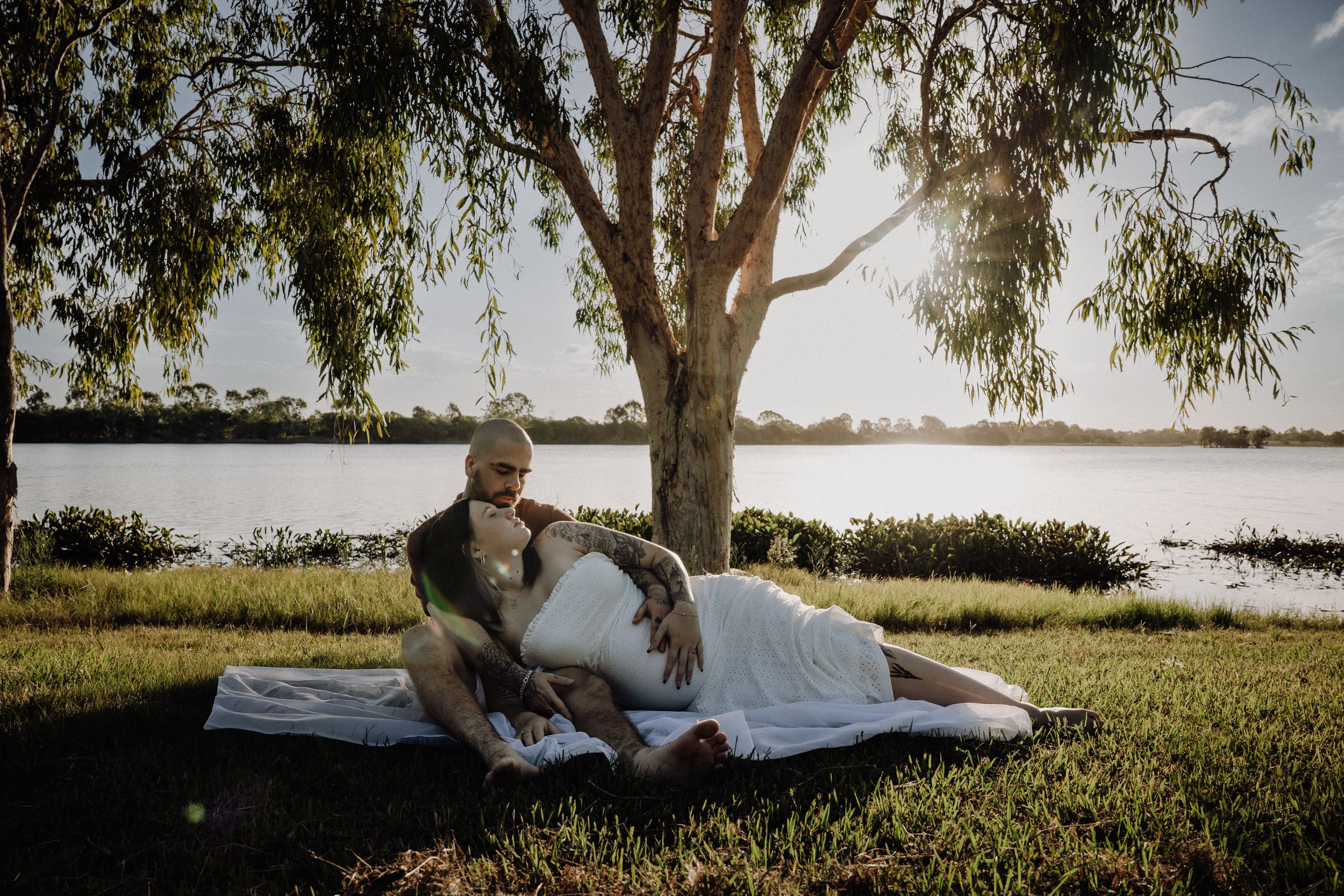 Couple in wedding attire relaxing on a blanket under a tree by a serene lake at sunset.