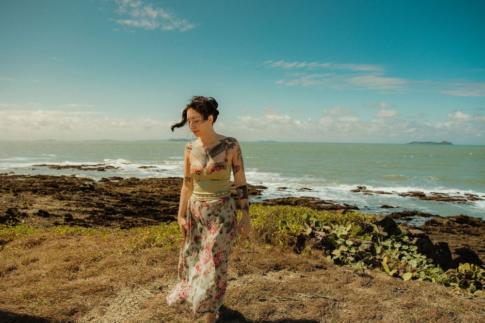 Woman in floral maxi dress standing on rocky coastal cliffs with turquoise ocean waves and blue sky behind her.