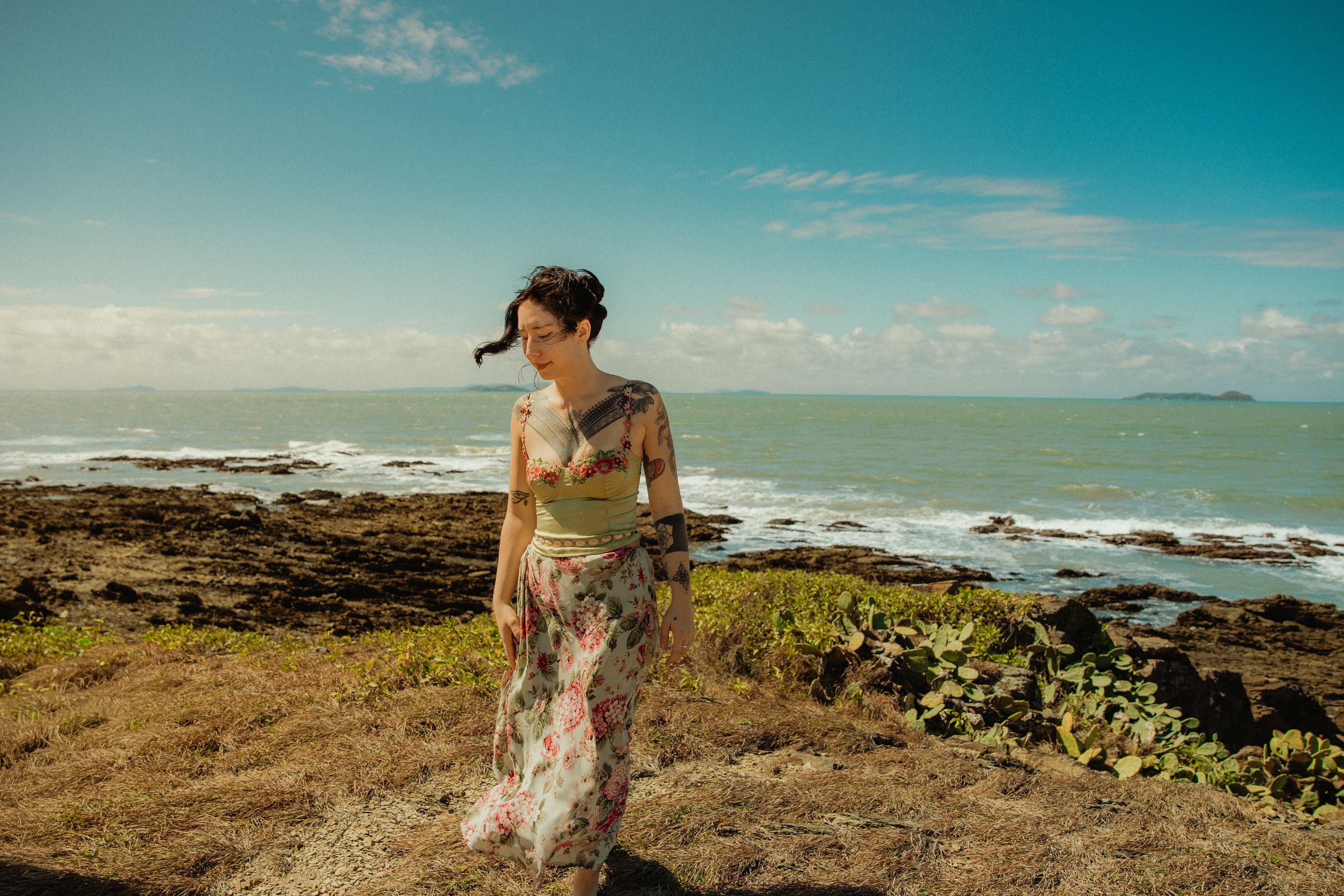 Woman in floral maxi dress standing on rocky coastal cliffs with turquoise ocean waves and blue sky behind her.