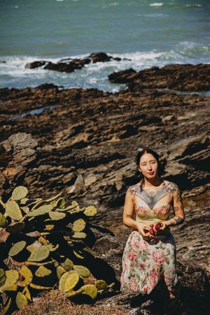 Woman in floral dress sits on rocky coastal shore beside cactus, holding flowers with ocean waves in background.