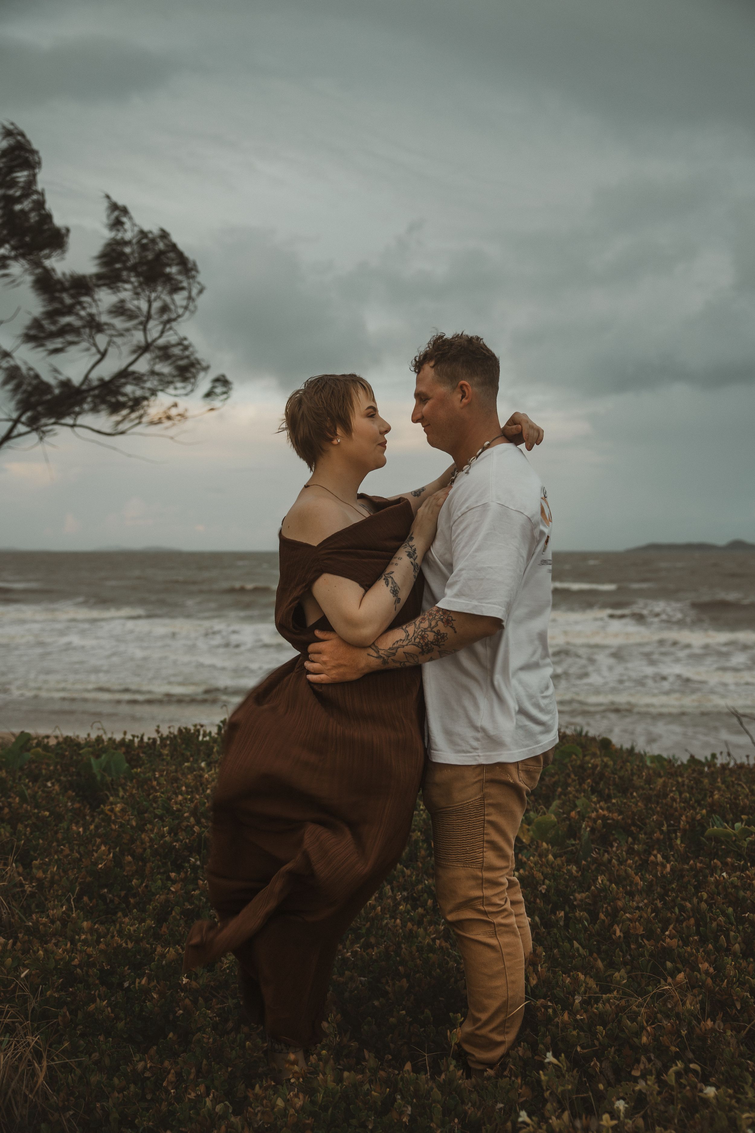 Couple embracing on a coastal cliff with stormy ocean waves, wearing brown dress and white shirt.