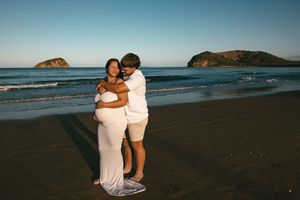 Couple embracing on a beach at sunset, pregnant woman in white dress, ocean waves and island in background.