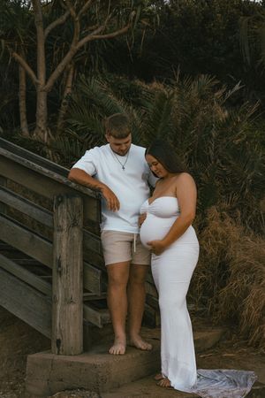 Couple poses on wooden steps outdoors, woman in white maternity gown cradling baby bump, man in white shirt and tan shorts.