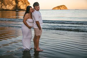 Pregnant couple standing in ocean waves at sunset on a beach, woman in white maternity gown, man in pink shirt.