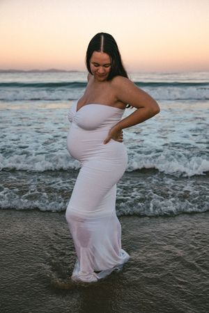 Pregnant woman in white wet dress standing in ocean waves at sunset beach maternity photo shoot.
