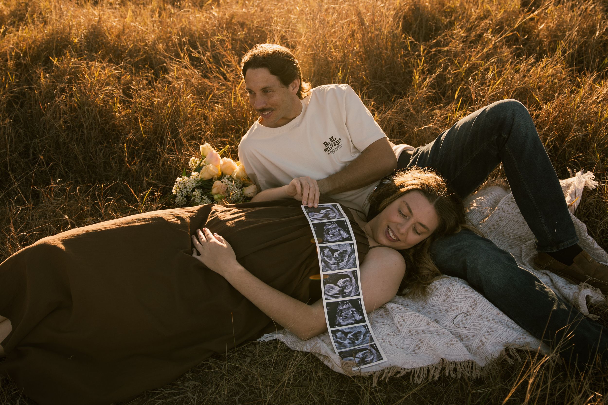 Couple sharing a tender maternity moment in golden field, woman lying down with ultrasound photos on her bump.
