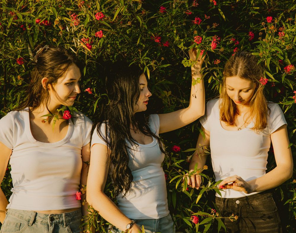 Three young women in white t-shirts pose together among lush green bushes with red flowers outdoors.