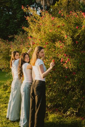 Three young women in white tops and wide-leg pants stand near a flowering bush in a sunlit garden.